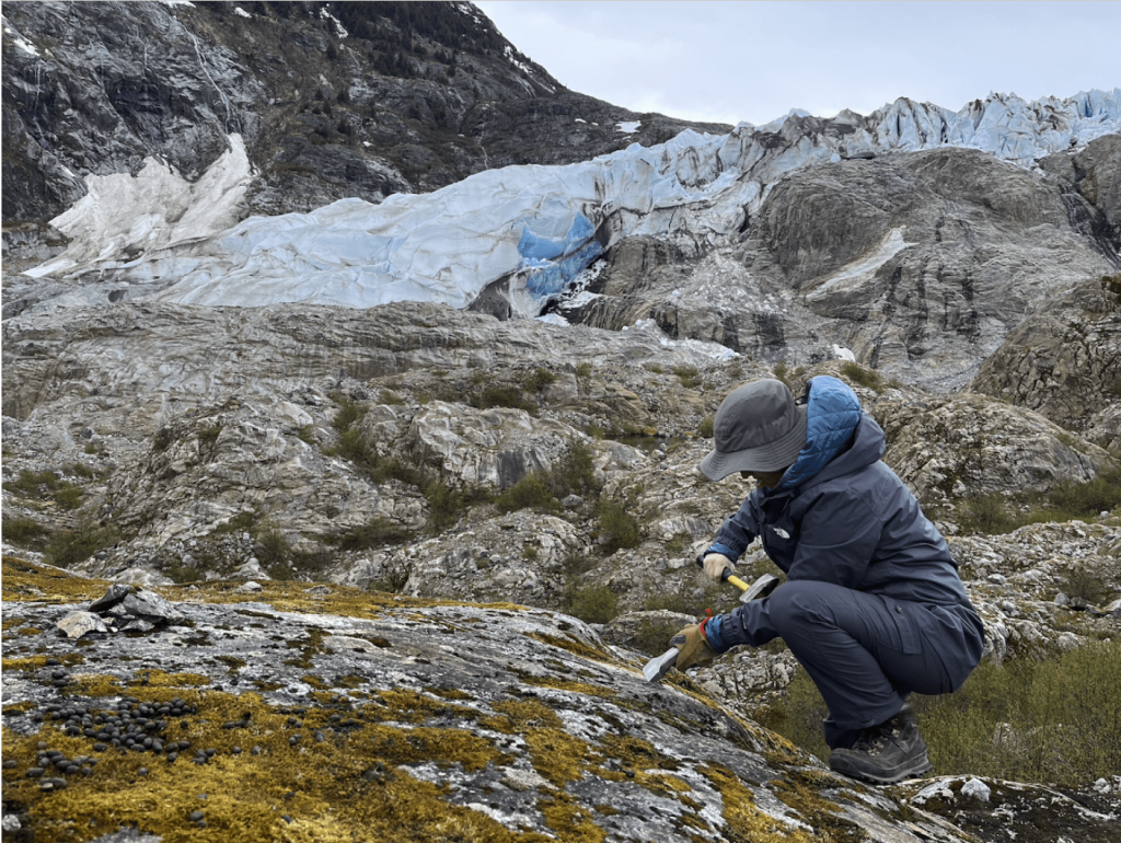 Student chiseling into a rock outcrop with a glacier in the background.