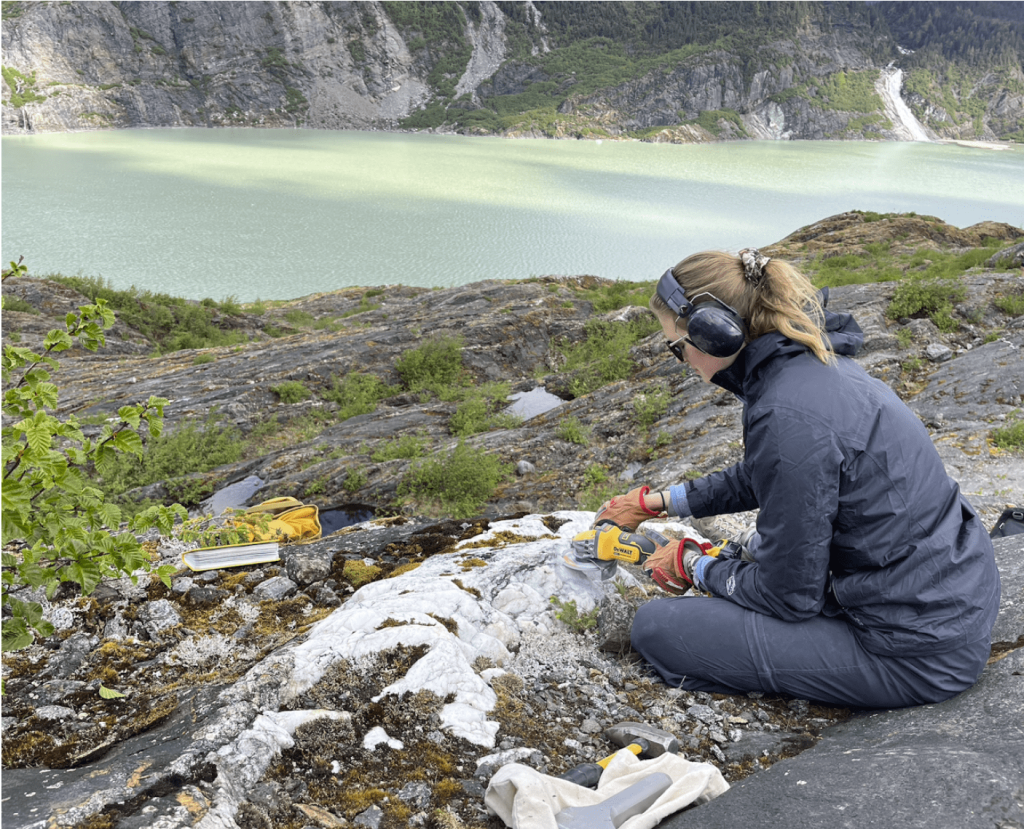 A student using a power tool to collect a rock sample with a view of the lake.