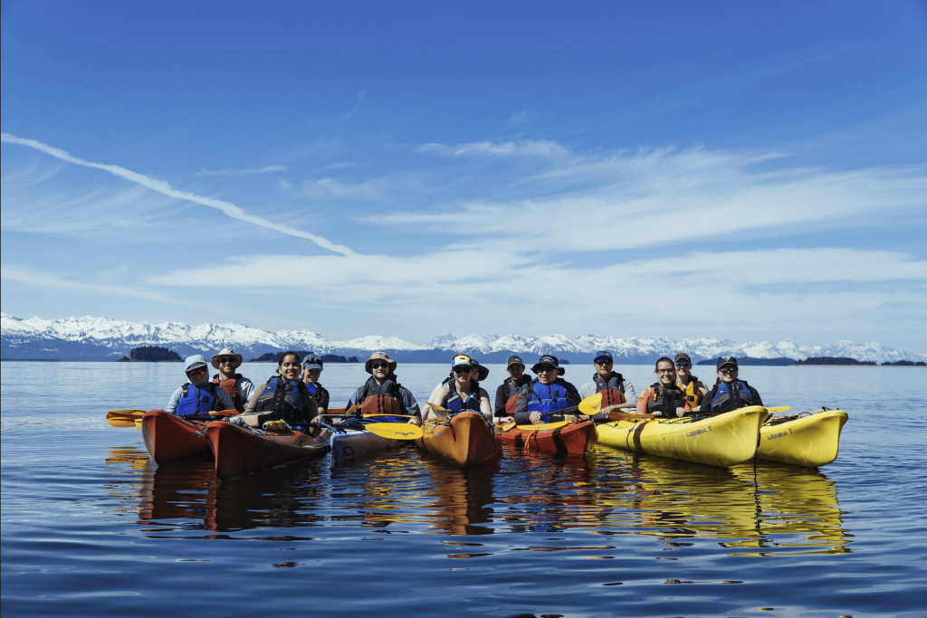 Student's in yellow and orange Kayak's in the lake smiling for the camera, mountains in the background