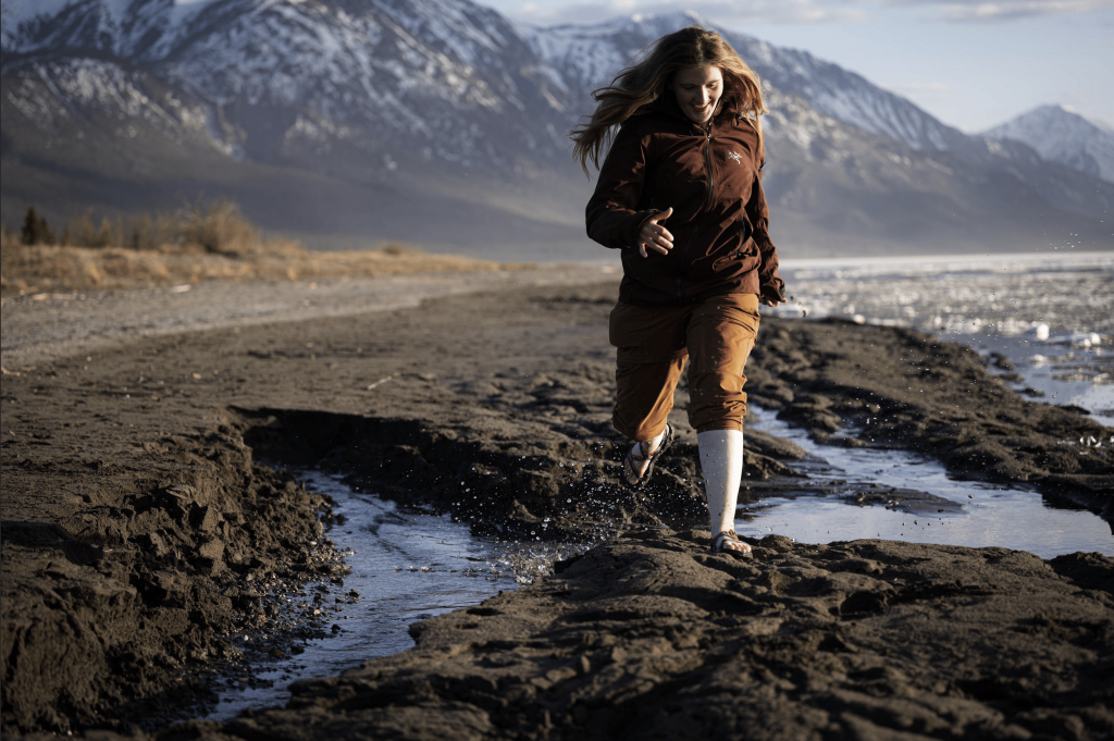 Student jumping across puddles on rocky shoreline with mountains in the background