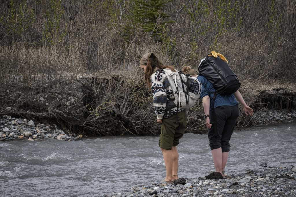 Two students standing barefoot in a rocky river with dense bushes in the background