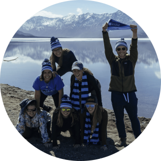 Six students stacked on top of each other in a pyramid while a fith student holds up a uMaine flag, with a lake and snowy mountains in the background.