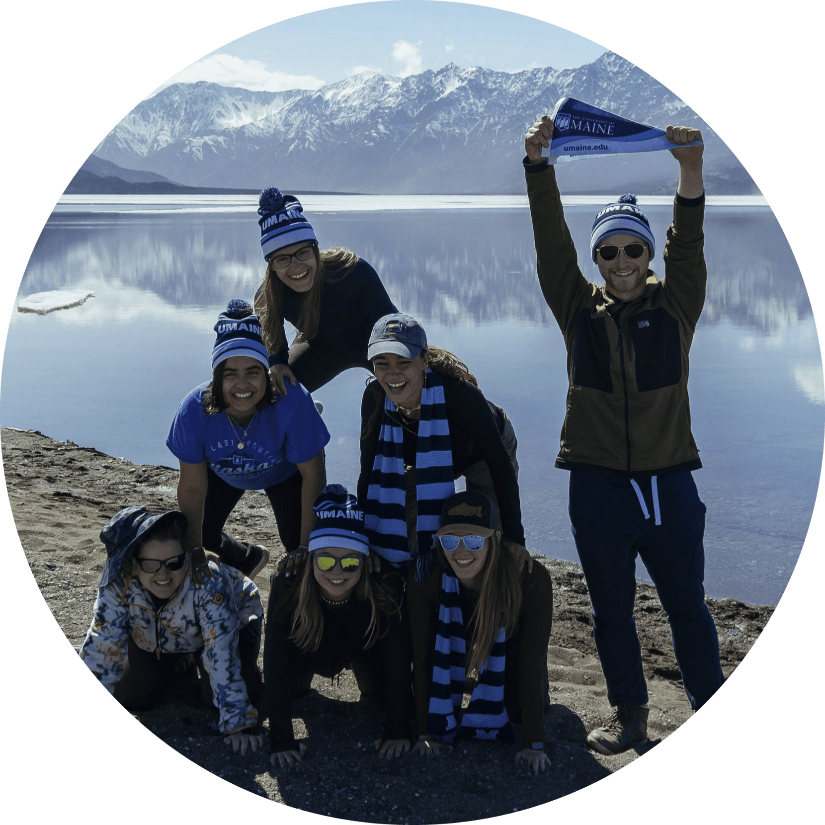 Six students stacked on top of each other in a pyramid while a fith student holds up a uMaine flag, with a lake and snowy mountains in the background.