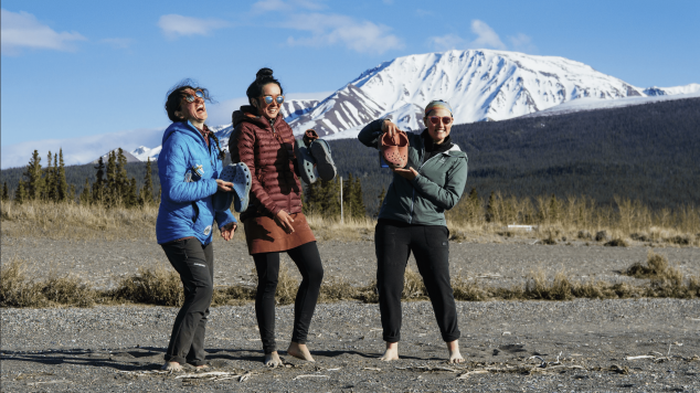 Three students laughing, holding Crocs, on a rocky field in front of a snowy mountain.