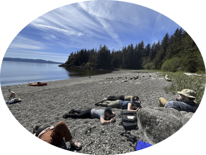 Students lounging on a rocky beach near a lake and forest.