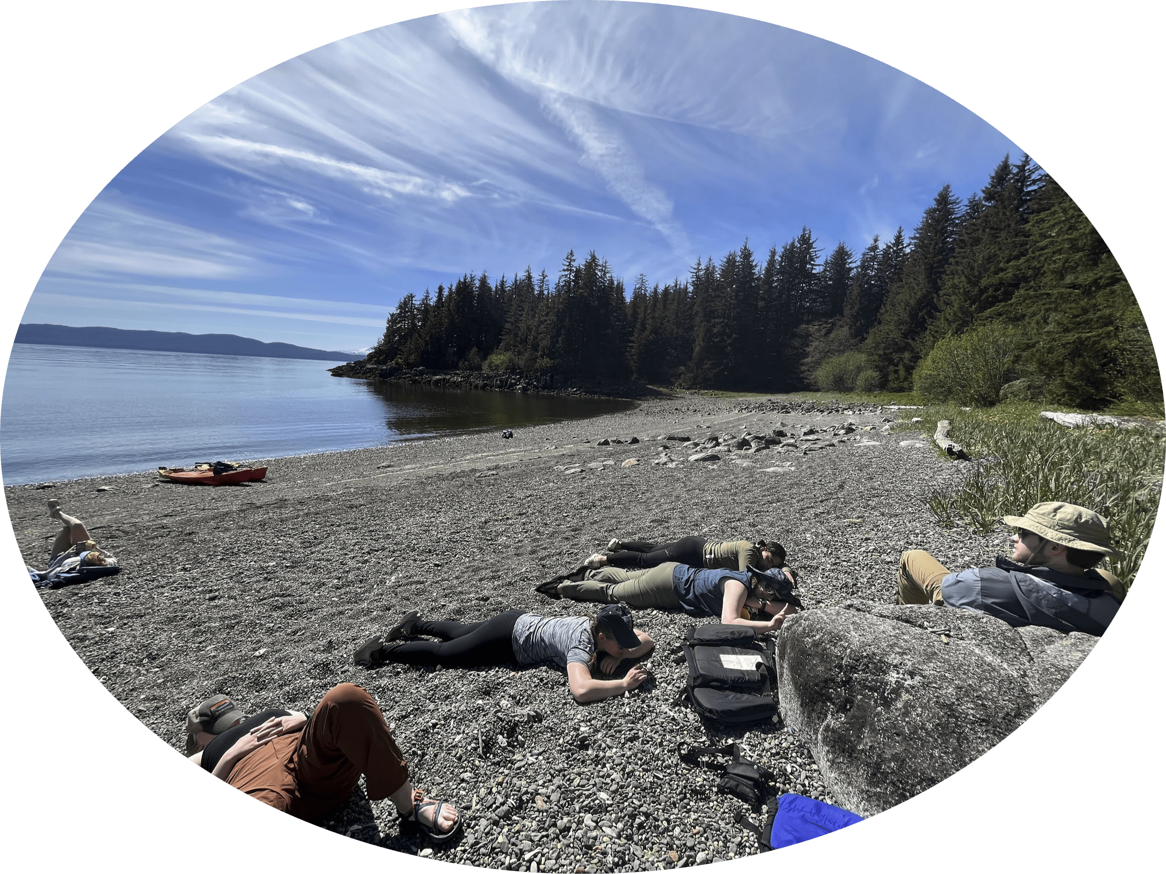 Students lounging on a rocky beach near a lake and forest.