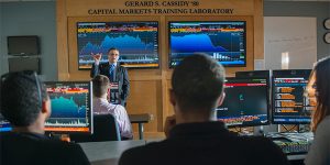 Students listening to a man teaching about the financial markets. Behind the teacher, there are two screens that display line charts of a stock.