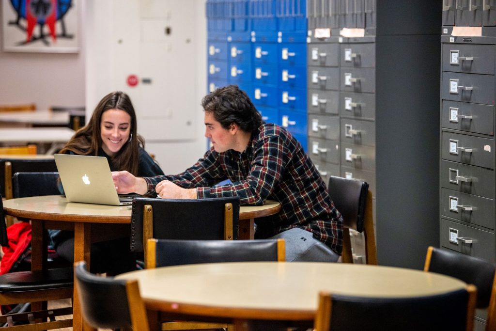 Two college students sit in the middle ground at a round table and look at a laptop. Behind them are metal filing cabinets.