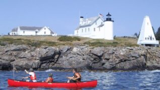 Three kids canoeing in front of a lighthouse at Blueberry Cove summer camp in St George Maine