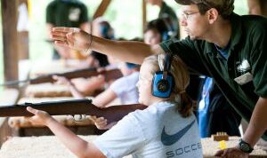 Male instructor helping female student shoot a rifle at a shhoting sports summer camp in Maine