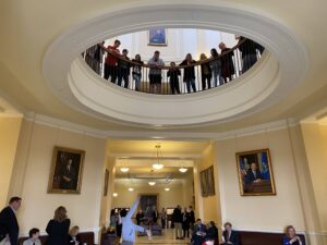 Students stand against a railed opening on the second floor at the Maine State House, they peer down into the first floor level.