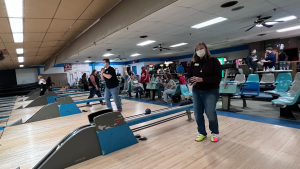 Several people in their own lanes preparing to roll a bowling ball, behind them are more people sitting and watching.