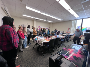 A group of students gather around a table in a 3D Printing Room at the University of Maine Augusta. Staff stands opposite of the students, explaining their work.