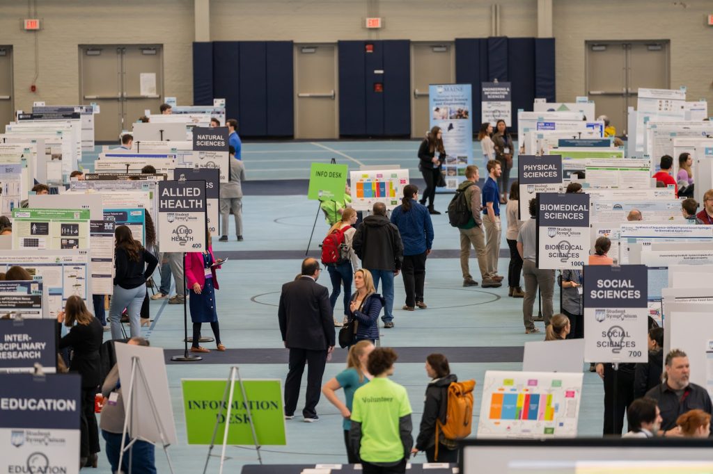 Wide shot of the middle aisle at UMSS24. Students, faculty, staff, and attendees fill teh aisle, looking at presentations.