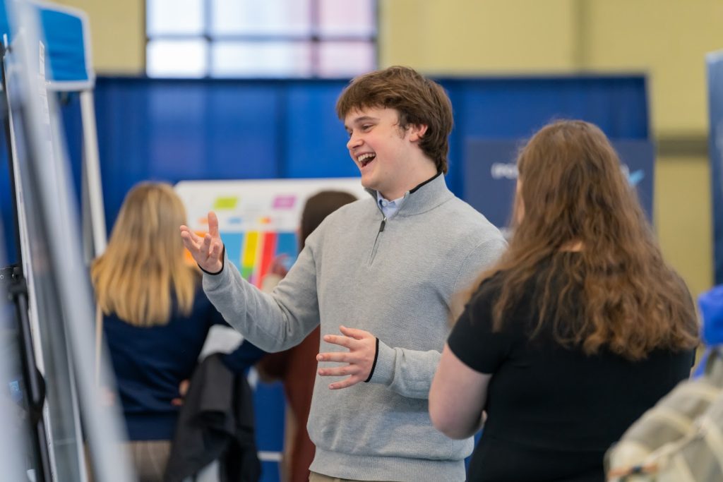 A student happily gestures to his poster as he explains his work to UMSS24 attendees.