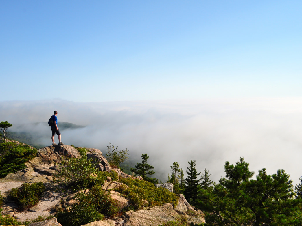 A hiker standing atop a mountain in Maine looking out on a cloud-swept vista.