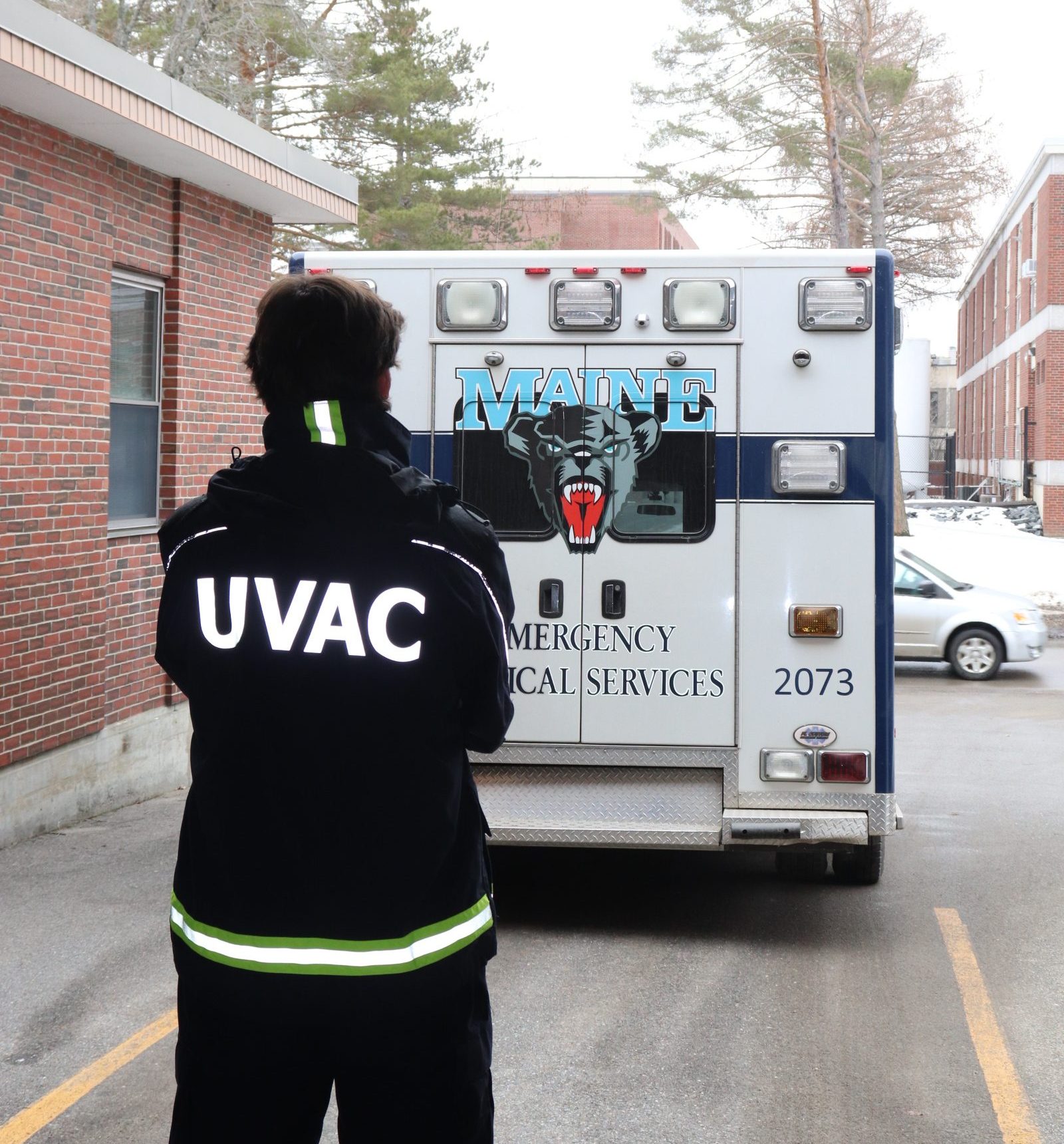 A UVAC member stands in the foreground facing away. He is wearing a jacket with "UVAC" on the back. In front of him our box-truck ambulance is parked on the ramp outside of the ambulance bay.