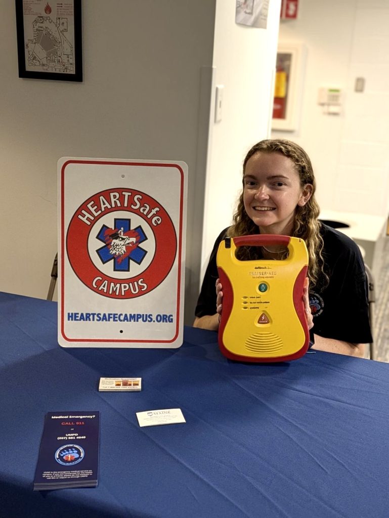 A UVAC member sits at a table directly behind and AED, with a Heartsafe Campus sign next to the AED.