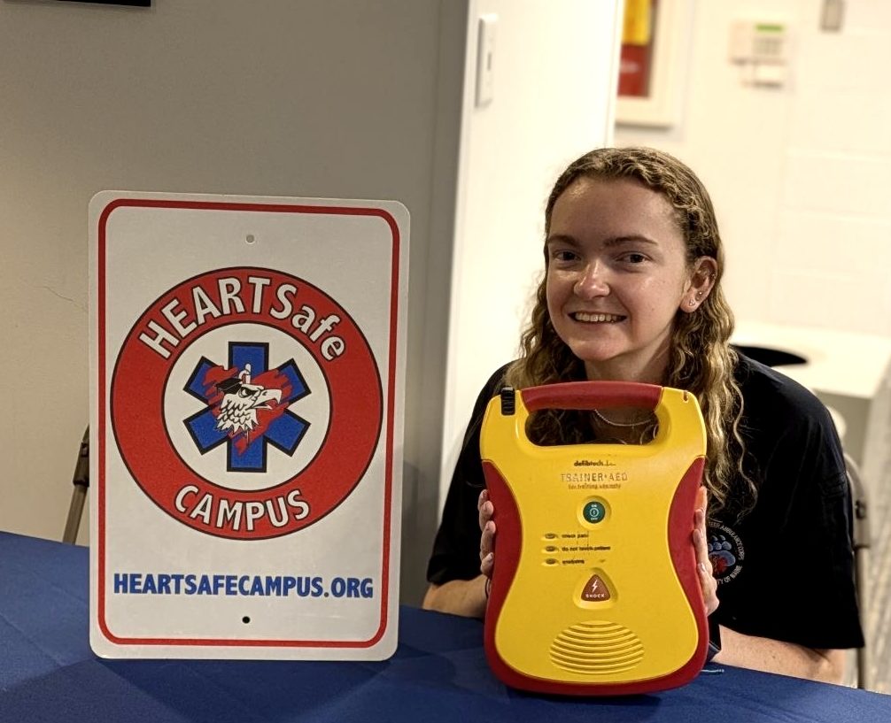 A UVAC member sits at a table directly behind and AED, with a Heartsafe Campus sign next to the AED.