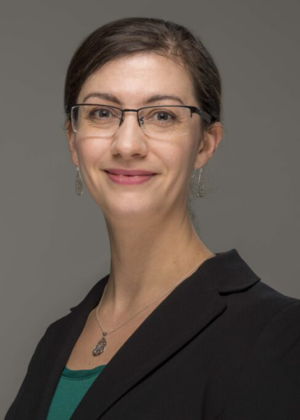 Dr. Caitlin Howell in a three-quarter pose, facing the camera. She is wearing a black blazer, green shirt, silver necklace, glasses, and is smiling at the camera.