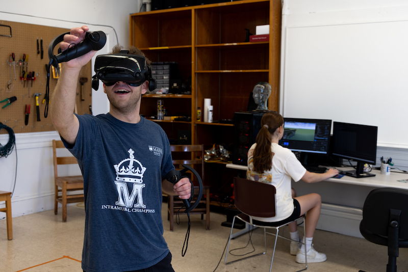 A male student in a VR headset reaches up with a controller while a female student works at a monitor in the background