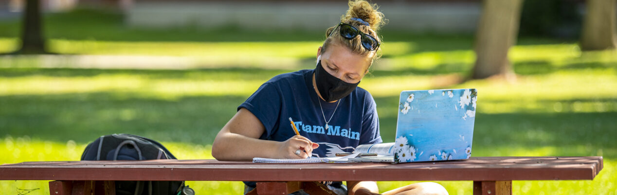 Student wearing face covering while working on laptop outside
