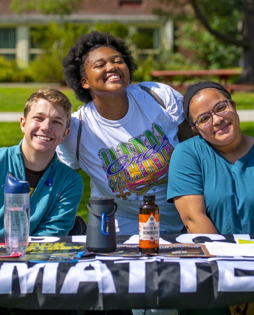 Three students smile while sitting at a table on UMaine's Mall