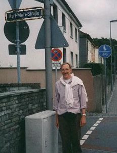 A man (younger Michael Howard) standing next to a European street named after Karl Marx.