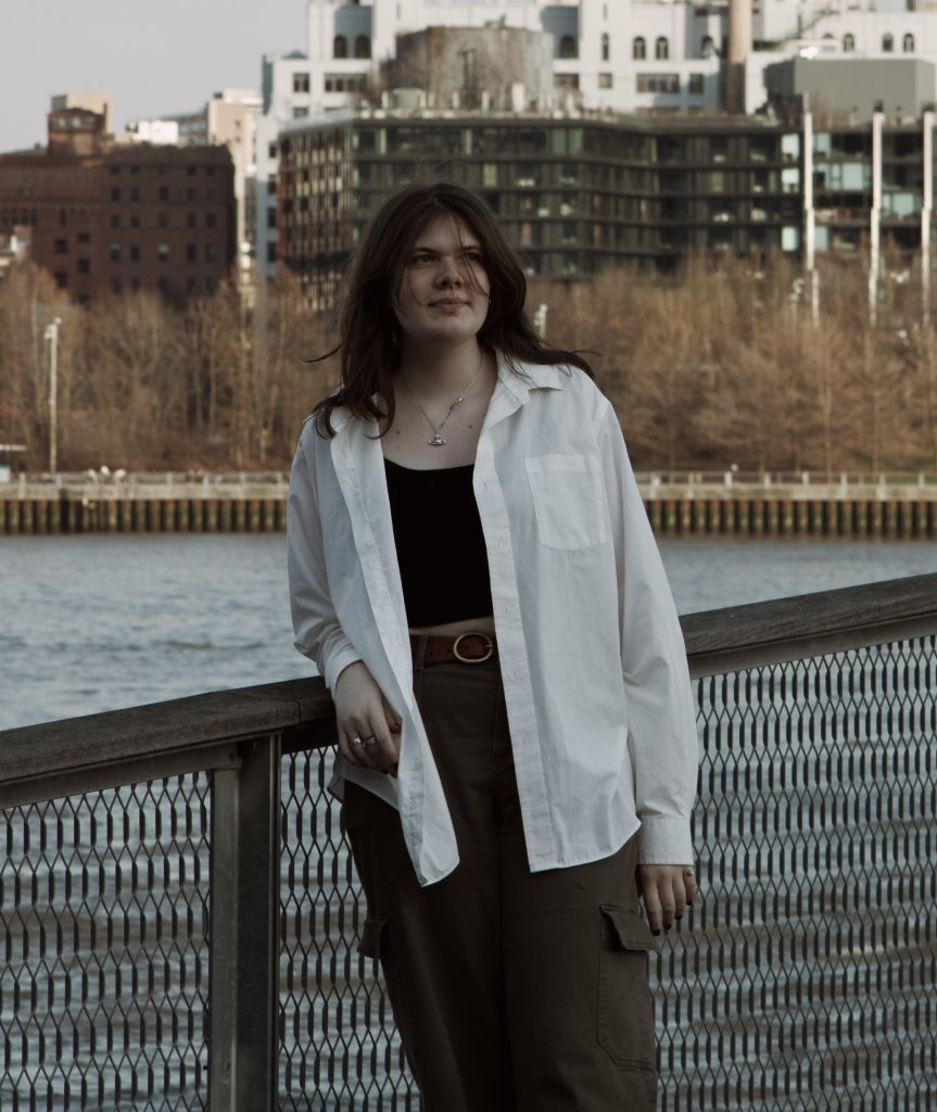 A girl (Maggie Kastelein) leaning over a fence with some coastal urban scenery in the background.