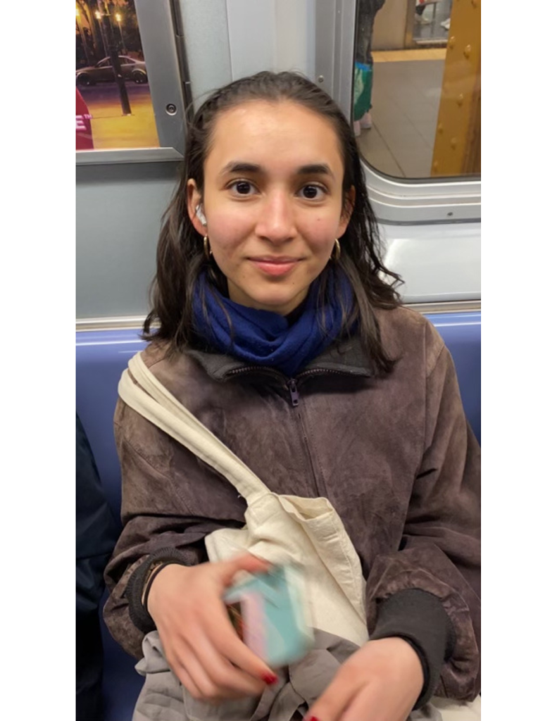 A girl (Rehannah Baksh) smiling wearing a scarf and tote bag around her sitting on a train or bus.