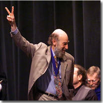 A man in a suit (Doug Allen) holding out a peace sign on a stage.